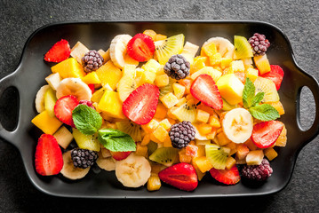 Vegan dietary foods. Vitamins. Dessert. Summer. Salad of fresh organic fruits mango, peach, apple, banana, kiwi, strawberry, blackberries. On black ceramic plate, black stone table Top view copy space