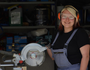 Fototapeta premium Junge lächelnde Frau im Overall mit Gehörschutz und Schutzbrille an Kreissäge