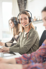 Portrait of cheerful young woman using telephone with headset and typing on a computer at her desk...