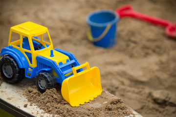 Closeup of bright blue and yellow tractor and children plastic shovel and pail in the sandbox. Baby's toys outdoor.