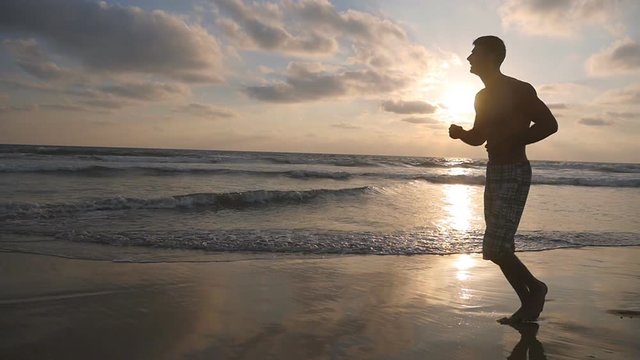 Athletic guy jogging along ocean shore during sunrise. Young sporty man running on the sea beach at sunset. Male sportsman exercising outdoor. Healthy active lifestyle. Workout at nature. Slow motion