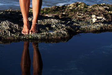 A young woman's toned legs and bare feet are reflected in a calm rock pool as she explores an...