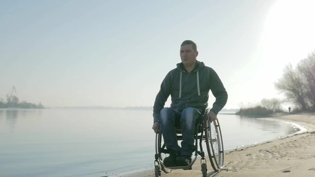 handicapped goes to embankment along sea on wheel chair, portrait lonely male cripple on wheelchair, alone man invalid outdoors, disabled on nature, faith in future, slow motion