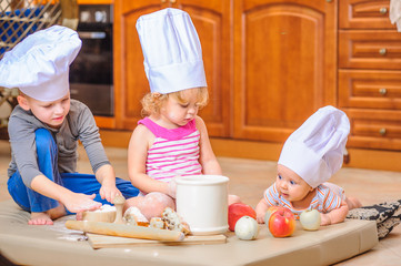 Two siblings - boy and girl - and a newborn kid with them in chef's hats sitting on the kitchen floor soiled with flour, playing with food, making mess and having fun