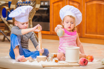Two siblings - boy and girl - in chef's hats sitting on the kitchen floor soiled with flour, playing with food, making mess and having fun