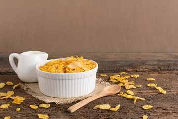 white bowl of cornflakes on sackcloth with wooden spoon and little pitcher of milk on wooden background with copy space ,still life , in concept breakfast