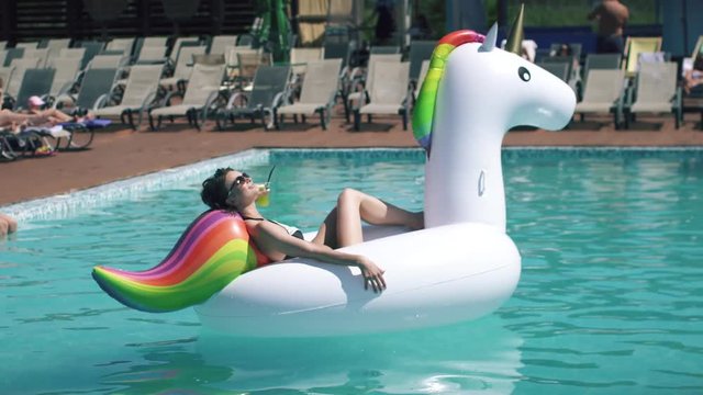 portrait of young sexy woman sunbathing on an inflatable mattress a unicorn in an outdoor pool. beautiful girl in sunglasses drinking a cocktail