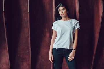 Attractive brunette woman in a white t-shirt stands on a dark wooden wall background. Mock-up.
