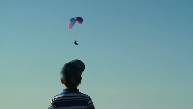 The boy is looking at a floating paraglider in the sky