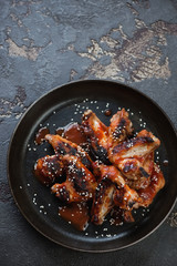 Above view of a metal plate with roasted chicken wings with sesame and tomato sauce, brown stone background with space