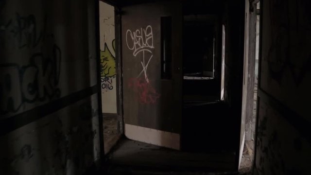 FPV, CLOSE UP: Walking along dark narrow hallway in abandoned decaying psychiatric hospital. Moving past the weathered wooden doors and broken crumbling walls in sinister sanatorium looking for escape