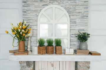 White wooden shelf with bouquet of tulips, grass in pot, greenery in vase, books and candle against stone wall with decorative window. Сlose-up decoration in living room at home