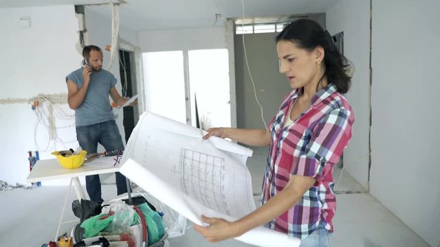Couple at their new home, woman reading blueprints, man working in the background
