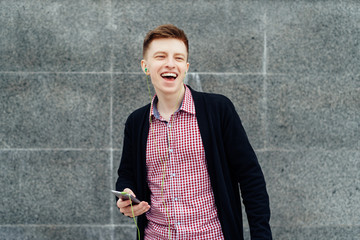 Cheerful happy young man in plaid shirt and jacket listening to music with headphones from cell phone and singing outdoors. Student with smart phone and headphones