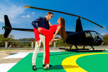 Naklejka premium Young elegant man in sunglasses and a woman in red standing next to a private helicopter.
