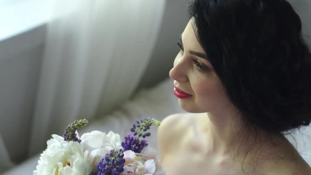 The morning of a gentle attractive brunette. Beautiful bride is smiling and looking out the window and a bouquet while sitting on the bed.