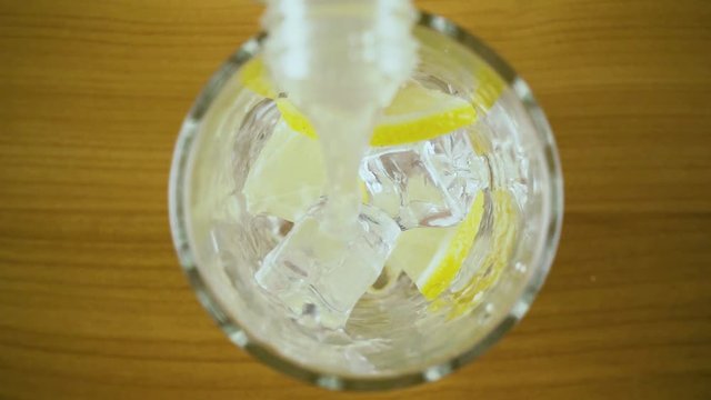 Lemon water from a bottle is poured into a glass with ice and lemon slices top view. Slow mo