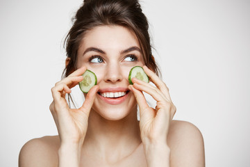 Young beautiful girl with perfect clean skin smiling holding cucumber slices over white background....