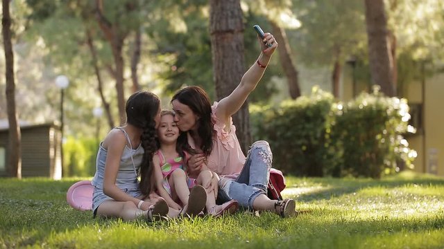 Mom with two daughters in the nature sitting on the grass and making a selfie with a phone