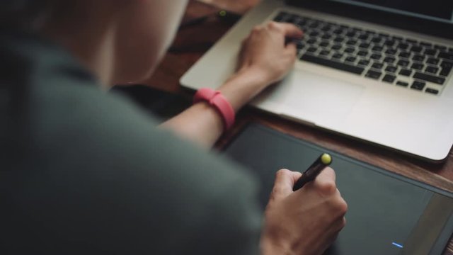 Woman designer working at a laptop with a graphics tablet. The view from behind shoulder, close-up