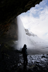 Person standing underneath cliff looking at view of waterfall, Iceland, Europe.