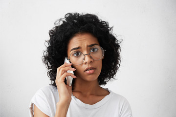 Close-up portrait of dark-skinned Afro American woman with dark abundant hair wearing big eyeglasses and casual T-shirt communicating over smartphone having sorrorful and sad expression. Emotions