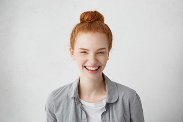 Portrait of red-haired woman with pleasant features dressed in casual grey shirt smiling with touching smile having fun indoors. Attractive lady with toothy smile posing in white studio over wall