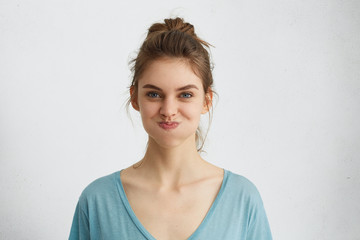 Indoor shot of blue-eyed pretty brunette woman making grimace blowing her cheeks having pleased expression. Beautiful young woman with pure healthy skin dressed in casual clothes being offended - obrazy, fototapety, plakaty