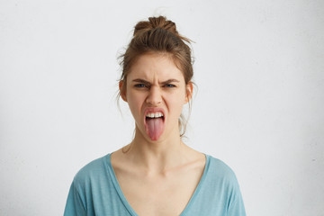 Grumpy woman with hair knot showing her tongue expressing negative emotions. Furious woman showing her disgust while having quarrel. Gloomy irritated blue-eyed female making grimace showing wrath © WHstudio Leushin N