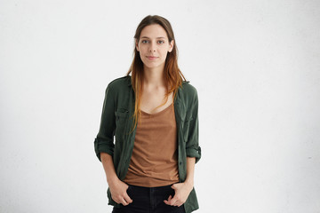 Horizontal portrait of beautiful woman with European appearane having oval face, dark attractive eyes and long straight hair dressed casually feeling relaxed while standing with hands in pockets