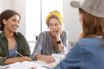 Exhausted bored woman with European appearane wearing yellow headband on head and casual shirt working with her groupmates together hearing her dark-skinned female friend writing notes in her notebook
