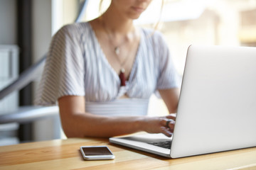 Cropped view of businesswoman sitting at wooden desk indoors surrounded with modern gadgets. Clever woman working online via laptop computer typing report. Woman's hands keyboarding on computer