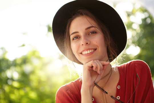 Charming young female with twinkling brown eyes wearing summer hat and red dress holding sunglasses looking pleasantly into camera. Young pretty girl with toothy smile admiring peace resting at nature