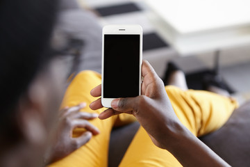 Back view of hipster guy lying at cozy sofa holding modern smartphone with blank screen for your text messaging and looking photos online. Cropped view of dark-skinned male holding mobile phone