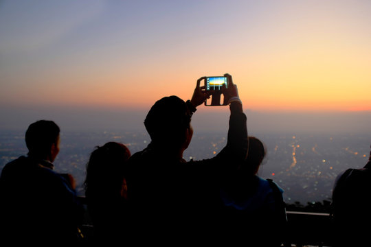 The image back side  of an Asian man using a mobile phone to take a picture of the morning scenery with the city lights as the background.
