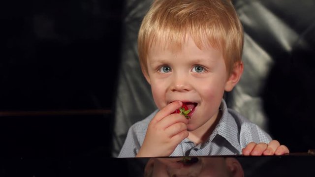 Little boy with blond hair eating a strawberry and showing thumbs up on a black background. 4K