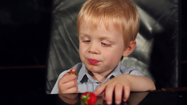 Close-up of a small child eating strawberries sitting at a table in a leather armchair on a black background.