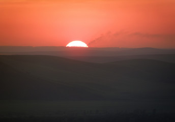 Large sun setting over the horizon in the hills covered with mist with smoking chimneys