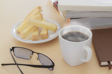Cup of coffee, biscuits and eyeglasses with books on table in morning.