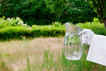 Hand of the waiter in a white glove and with a white napkin holding an empty glass decanter on a blurred background of nature green bushes and trees