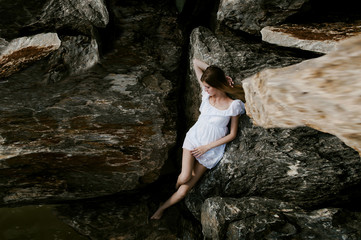 Portrait of slender young woman on stones near the sea. Beautiful girl is resting on coast, enjoying outdoor recreation. Lies on rocks, sexy white dress, graceful long thin tender hands and feet