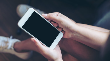 Mockup image of woman's hand holding white mobile phone with blank black screen on thigh with wooden floor background in modern