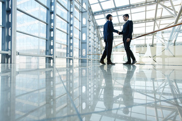 Wide angle portrait of two contemporary business men shaking hands in hall of futuristic glass building