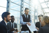 Portrait of modern businessman presenting contract documentation to group of people at table in bright sunlight