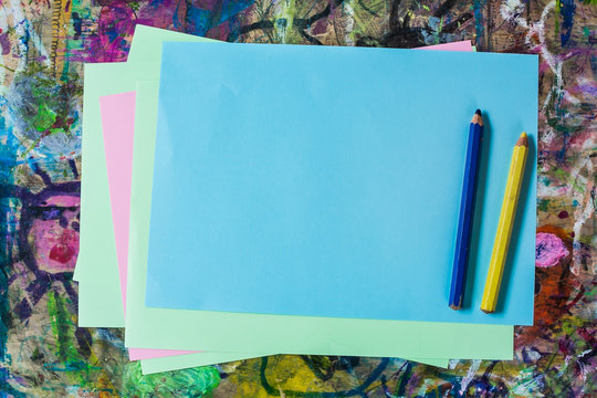 Colorful sheets of paper and two pencils, yellow and blue, on kid's painting table dabbled with random stains of paint - horizontal.