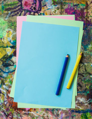 Colorful sheets of paper and two pencils, yellow and blue, on kid's painting table dabbled with random stains of paint - vertical.