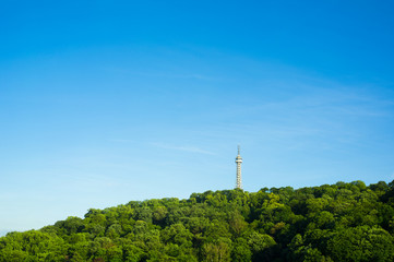Fototapeta na wymiar Petrin Lookout Tower ( Petrinska rozhledna ), Prague, Czech Republic / Czechia - tall building on the top of the hill. Green trees around it. Minimalist composition with blue sky as copy space