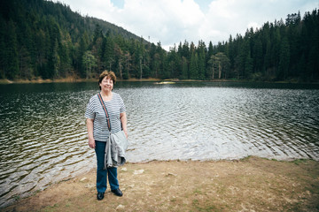 Fototapeta na wymiar Aged beautiful woman standing on the bank of the mountain lake surrounded by forest. Travel concept