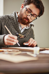 Bearded painter in eyeglasses and sweater drawing by workplace