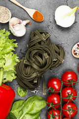 Raw pasta of tagliatelle with spinach and ingredients for cooking (cherry tomatoes, spices, garlic, spinach, pepper, lettuce) on a dark background. Top view. Food background